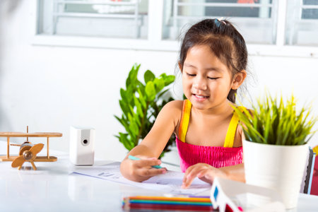 Child little girl drawing cartoon on paper before paint the color, Asian cute kid preschooler sitting on table smiling she draw and writing a picture with pencil at home to learning arts homeworkの写真素材