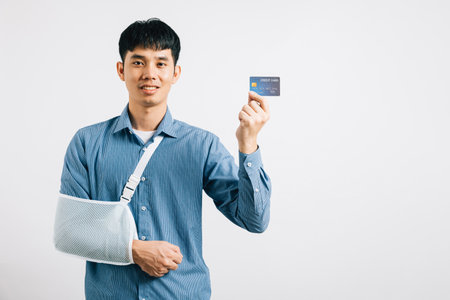 Despite a damaged arm, a man wears a support splint, paying medical bills with a credit card after an accident. Happy Asian man in a sling on white background, emphasizing health care. Copy spaceの写真素材
