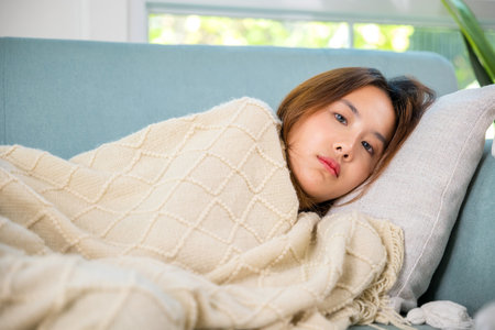 Asian sick woman cold high fever covered with blanket resting sleeping at home, young woman have problem with headache lying on sofa in living room, Unhappy sick girl, person tiredの写真素材