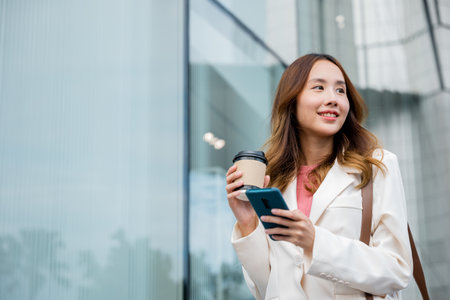 Young Asian freelance businesswoman looking away with smile while using smartphone and holding coffee cup on the go outdoors in urban park in the city. Remote working concept with flexible lifestyle.の写真素材