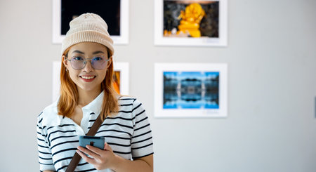 Visitor woman smiling at picture art gallery collection in front framed paintings pictures she holding mobile phone, Asian female watching at photo frame with smartphone at artwork gallery showの写真素材