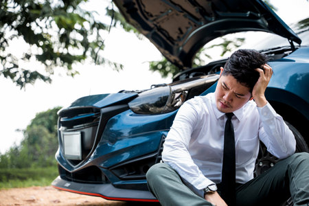 Man sitting near his broken car with raised hood on rural road, calling for roadside assistance. Concept of car trouble and frustration.の写真素材