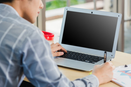 Asian businessman writing notes with pen inspired ideas list on paper notepad while working on laptop computer. Happy handsome man sitting busy on desk table working from home office doing noteの写真素材