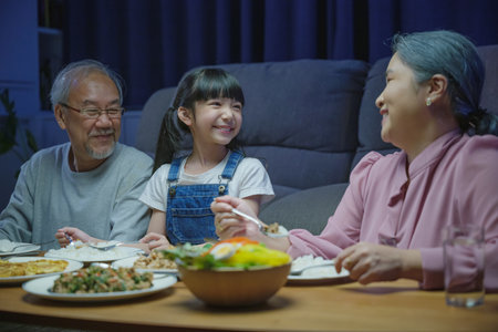 Happy Asian family grandparent and granddaughter dining on table and having fun during at house evening time, Happiness senior parent and child eating dinner food together in living room indoorsの写真素材