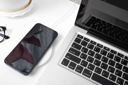 Charging smartphone battery with wireless charger device near laptop computer, Modern technology transfer energy mobile phone on workspace desktop at home officeの写真素材