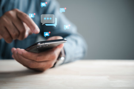 Home office setup man engages smartphone, typing for live chat and social network concepts. Chat box icons emerge, symbolizing digital communication and social media marketing.の写真素材
