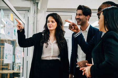 A group of professionals, led by a young businesswoman, discuss ideas and strategies in a meeting by a glass wall. Their teamwork, brainstorming, and creative thinking drive success.の写真素材