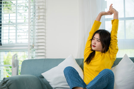 Happy woman stretch oneself for relaxing activity feeling lazy on weekend on couch, resting napping break, Lifestyle Asian young female stretching her arms on sofa in the living room at homeの写真素材