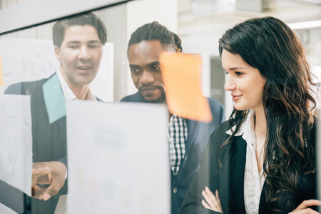 A diverse group of professionals meets in the office, using post-it notes on a glass board to...