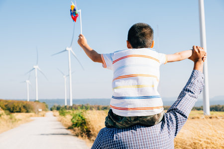 Father smiles carrying daughter with pinwheel. Family joy near windmills embodies global innovation in renewable energy. A bonding moment filled with happiness and community. Father Dayの写真素材