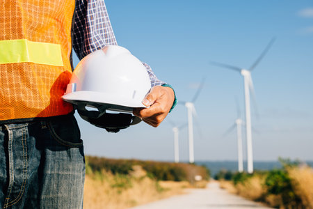 Man engineer in uniform holds helmet at windmill site. Symbolizes renewable success innovation tackling global warming. Leadership safety commitment evident in industrys progress.の写真素材