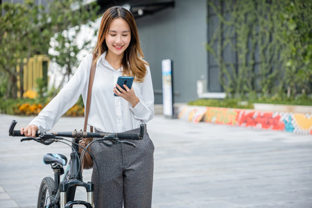 Asian businesswoman standing on city street building with bicycle holding smart mobile phone, lifestyle smiling young woman commute with her bicycle and use smartphone at urban for work social mediaの写真素材