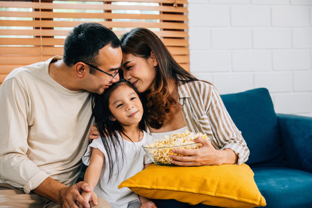 Family bliss is captured as they gather on sofa watching TV and munching popcorn. father mother daughter and sibling share laughter embodying happiness togetherness and relaxation.の写真素材
