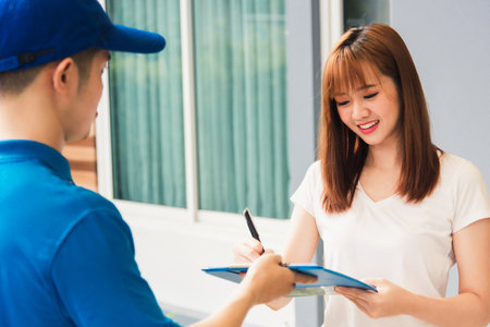 Asian beautiful customer young woman signing delivery in clipboard get her package from service courier man, smiling female signature receipt of delivery package at door front houseの写真素材
