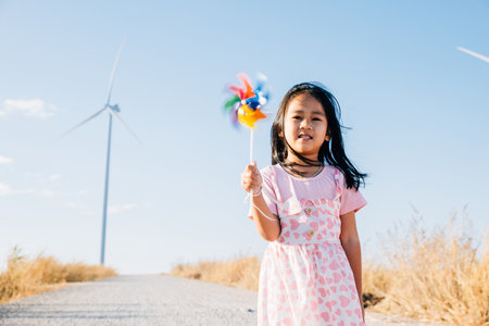 A joyful little girl smiling and running with pinwheels near windmills. Embracing clean energy through playful education in a picturesque wind turbine setting under a beautiful sky.の写真素材
