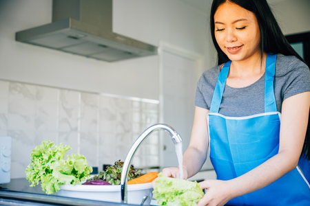 A smiling woman washes vegetables in the kitchen sink readying them for a delicious salad. Emphasizing hygiene and her passion for healthy eating in her home.の写真素材