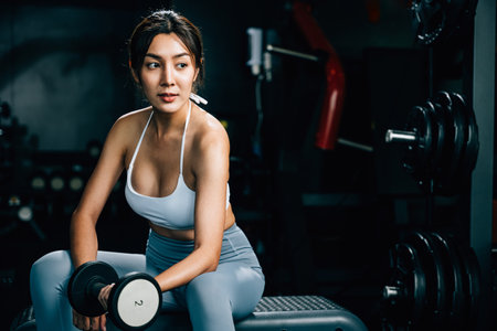 A sporty young woman sitting on a bench at the gym, practicing weightlifting with dumbbells to lift her bicep muscles and improve her physical strength and health, fitness GYM dark backgroundの写真素材