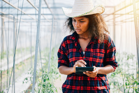 In a greenhouse a smiling woman farmer uses her phone to carefully inspect tomato leaves. Her modern approach to farming balances technology and nature emphasizing growth and industry development.の写真素材