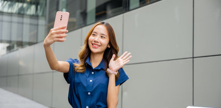 Smiling young lady taking a selfie with her cell phone in the city, showcasing her trendy style and...