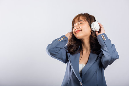 A cheerful woman with wireless headphones smiles while listening to music on her phone. Studio shot, isolated on white. This happy Asian young female is captivated by modern technology.の写真素材