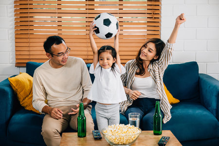 A jubilant family, with popcorn and a ball, raises their arms, cheering while watching a football match at home. Their togetherness and excitement reflect the joy of winning.の写真素材