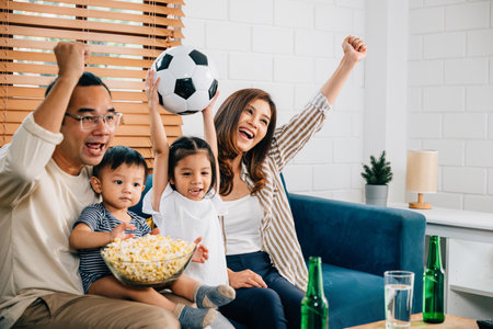 Delighted parents and children enjoy a football match at home, celebrating a goal with popcorn and a ball. Their excitement and togetherness capture the joy of winning and family fun.の写真素材