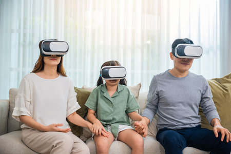 Asian family wear vr glasses headsets sitting on sofa in living room, game entertainment innovation technology, Happy family have mother father and daughter watching movie or playing video gameの写真素材