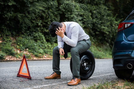 Expertise and skill in action as a young businessman in white shirt changes the wheel of broken car, while worried mature man looks on. A red triangle sign signals caution to other drivers on the roadの写真素材