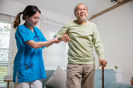 International Day for the Elderly. Young carer supporting helping senior man stand up with walking stick from sofa, Nurse wearing blue uniform help helping his patient senior old man with walking caneの写真素材