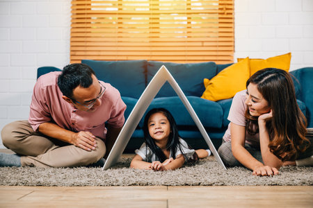 A happy family on a couch holding a cardboard roof in their new home symbolizing safety and support. Asian parents and their daughter embody affectionate togetherness.の写真素材