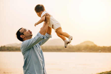 A happy dad and his toddler son share a playful moment of freedom and joy in the park, throwing him up in the air on a sunny summer day. Family love and happiness captured in a photographの写真素材