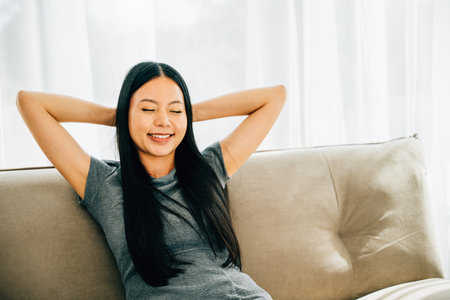 Relaxed woman on sofa hands behind head enjoys serene moment. Dreaming finding balance in modern living room. Embracing relaxation wellbeing and mindfulness at home. Kick back and relax conceptの写真素材