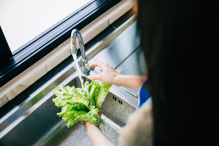 In a modern kitchen a womans hands wash fresh vegetables under running water in the sink for making a vegan salad. Cleanliness and freshness in homemade healthy food prep.の写真素材