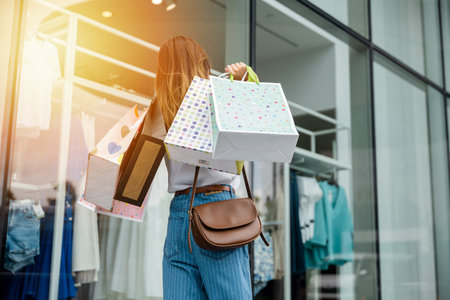 Back view of young woman holding shopping bags and walking in front of a clothing store, enjoying a summer day of retail therapy. She is a trendy consumer, always looking for the latest fashion items.の写真素材