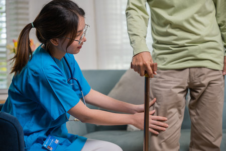 Asian young woman nurse checking knee and leg after surgery of senior old man patient suffering from pain in knee, doctor asking elderly man about pain symptom with walking stickの写真素材