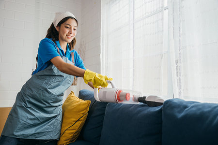 In a comfortable living room an Asian woman maid uses a vacuum machine cleaner to clean a sofa. Her dedication to hygiene and furniture care reflects modern approaches to housework.の写真素材