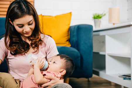 A heartwarming family portrait comes to life in the living room at home as a smiling mother holds her cute Asian baby who is feeding from a baby bottle radiating joy and togetherness in every glance.の写真素材