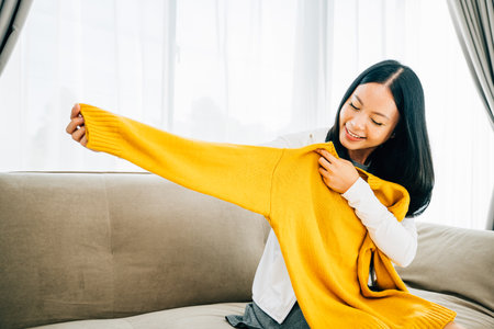 Happy Asian woman reveals a shirt from a box smiling with delight at home. Satisfied shopper unboxes...