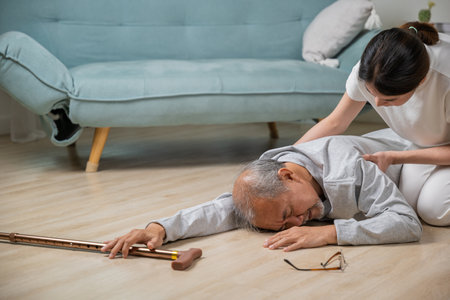 Dizziness elderly old man falling down lying on floor with walking stick and assistant nurse help to support, senior father fall on the ground and his daughter helping wake up and get up, accidentの写真素材
