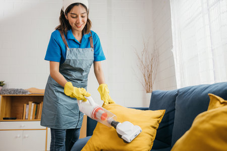 Focused on housework an Asian woman maid cheerfully vacuum machine cleaners a sofa in a living room....