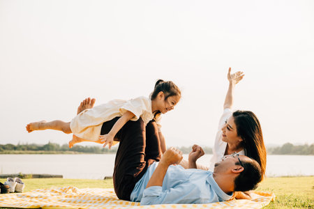 A joyful family moment in the park. Father holds his daughter up high as she smiles and flies like an airplane, while mother looks on with delight. Family fun at its bestの写真素材