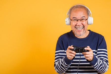 Portrait Asian smiling old man with headphones holding game controller studio shot isolated yellow...