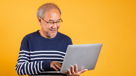 Portrait Asian old man wearing glasses use laptop computer studio shot isolated yellow background,...