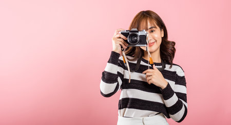 Attractive energetic happy Asian portrait beautiful young woman smiling photographer taking a picture and looking viewfinder on retro vintage photo camera ready to shoot isolated on pink backgroundの写真素材