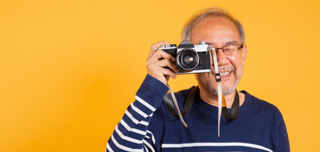 Portrait Asian old man wearing glasses look viewfinder vintage SLR camera studio shot isolated yellow background, smiling happy Photographer elderly man gray haired taking a picture of himselfの写真素材
