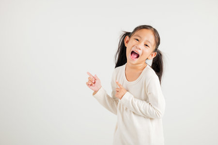 Happy Asian portrait cute young kid girl smiling indicate finger empty space, studio shot isolated on white background, Thai kindergarten child pointing index out with copy spaceの写真素材