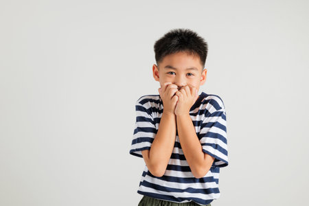 Asian portrait cute young kid boy standing amazed, shocked afraid mouth covered gesturing hand palms looking camera, studio shot isolated on white background, Thai primary child scaredの写真素材