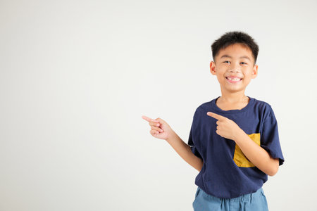 Happy Asian portrait cute young kid boy smiling indicate finger empty space, studio shot isolated on white background, Thai primary child pointing index out with copy spaceの写真素材
