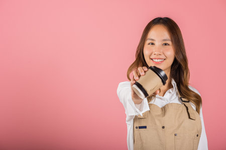 Portrait of Asian happy young woman barista bar tender coffee maker holding coffee tea hot cup studio shot isolated on pink background, female person smiling hold takeaways cup, small businessの写真素材