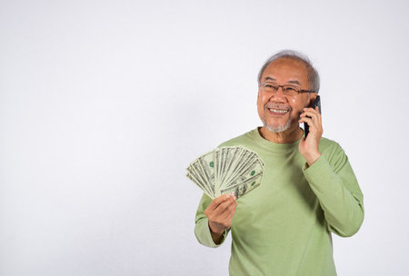 Portrait Asian smiling old man is holding a bunch of money fan and talking on cell mobile phone...
